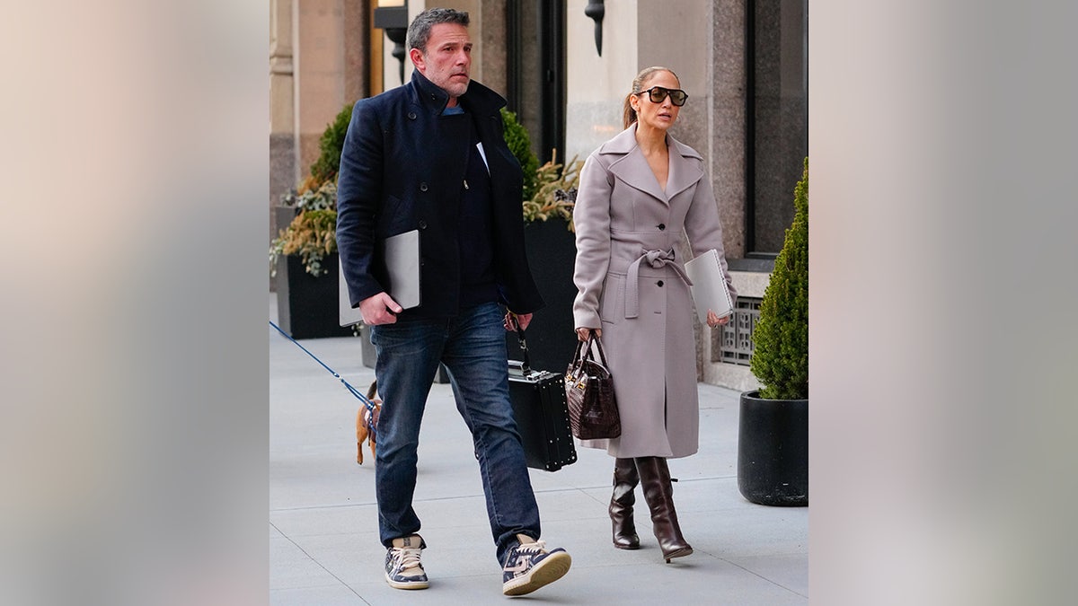 Ben Affleck in a pea coat and jeans walks with his computer in one arm and a briefcase in the other with Jennifer Lopez in a gray coat