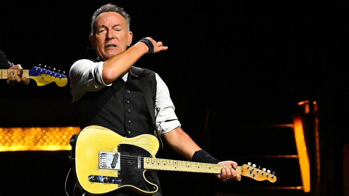 Bruce Springsteen performs on stage in Pittsburgh, wearing a black vest and playing a yellow guitar, swiping his hand in the air