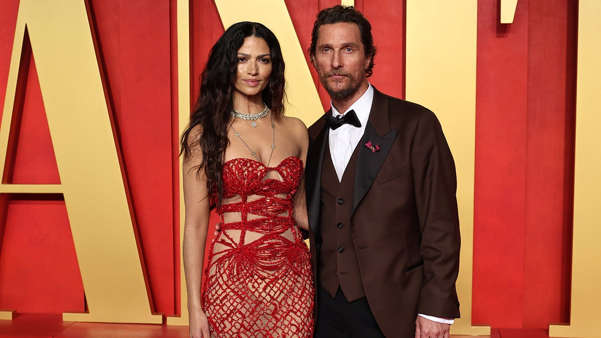 Camila McConaughey in a red beaded and wired strapless dress poses with husband Matthew McConaughey in a dark brown/burgundy suit at the Vanity Fair after party
