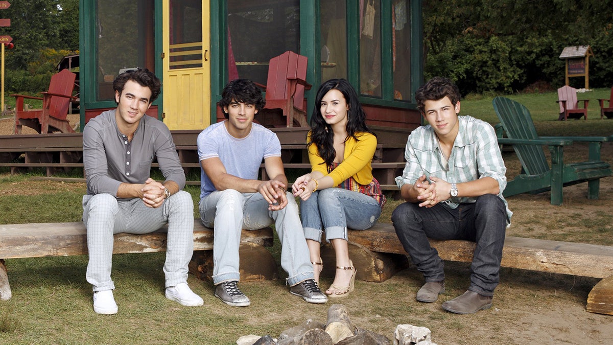 Demi Lovato in a yellow shirt sits on a wooden bench with the Jonas Brothers for 