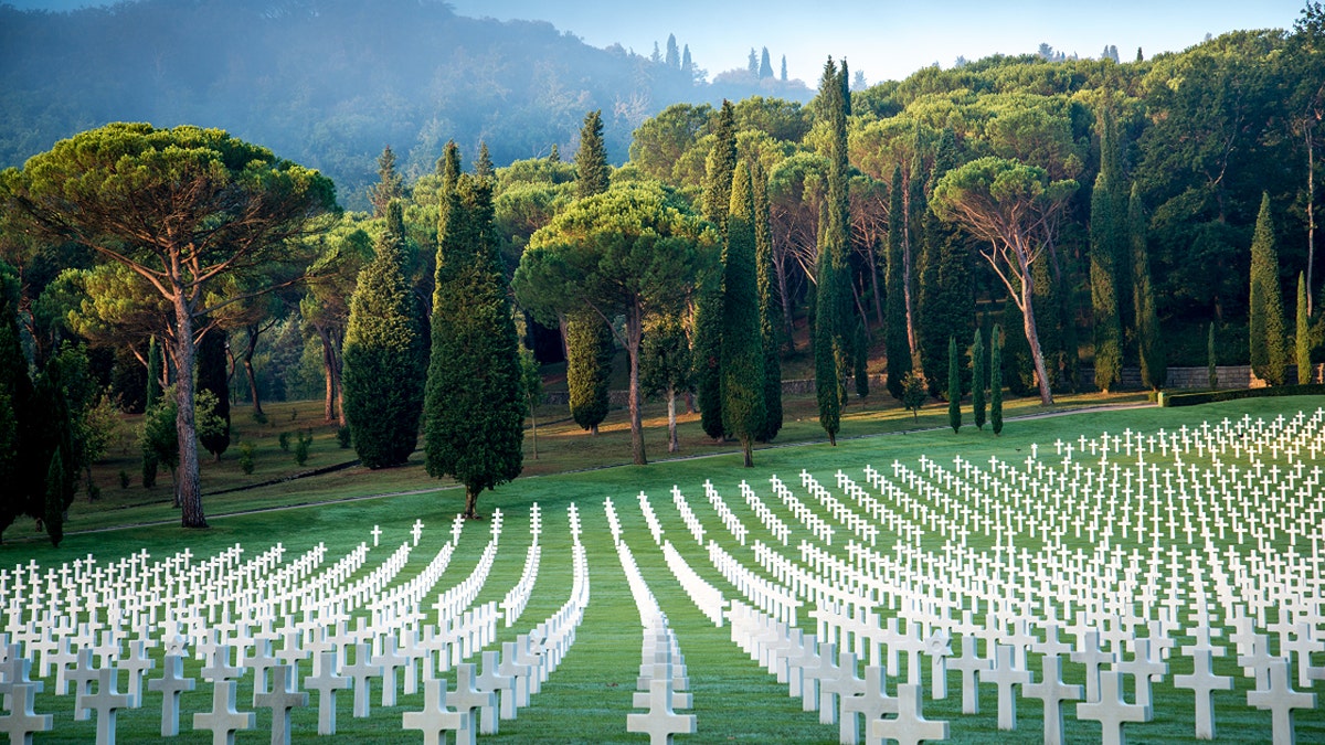 Rows of cross-shaped headstones extend towards lush trees in the Florence American Cemetery in Italy with mountains in the background.