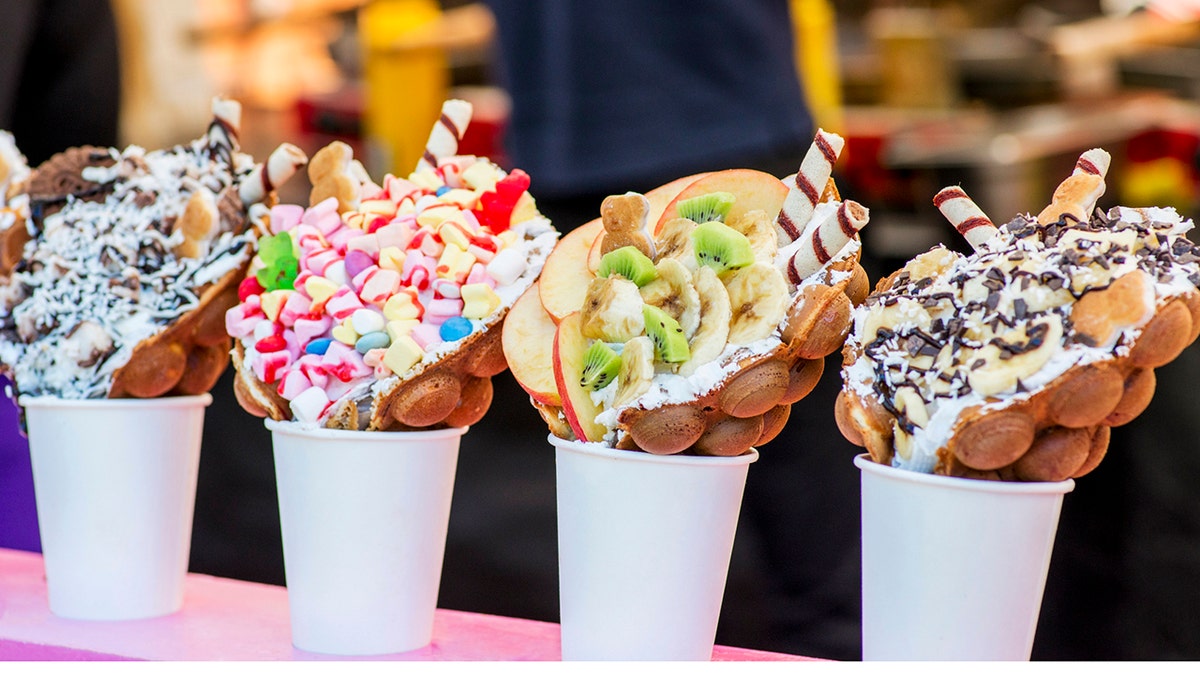 Bubble waffles with raspberries, chocolate and marshmallow in white paper cups are sold on Open kitchen food festival event.