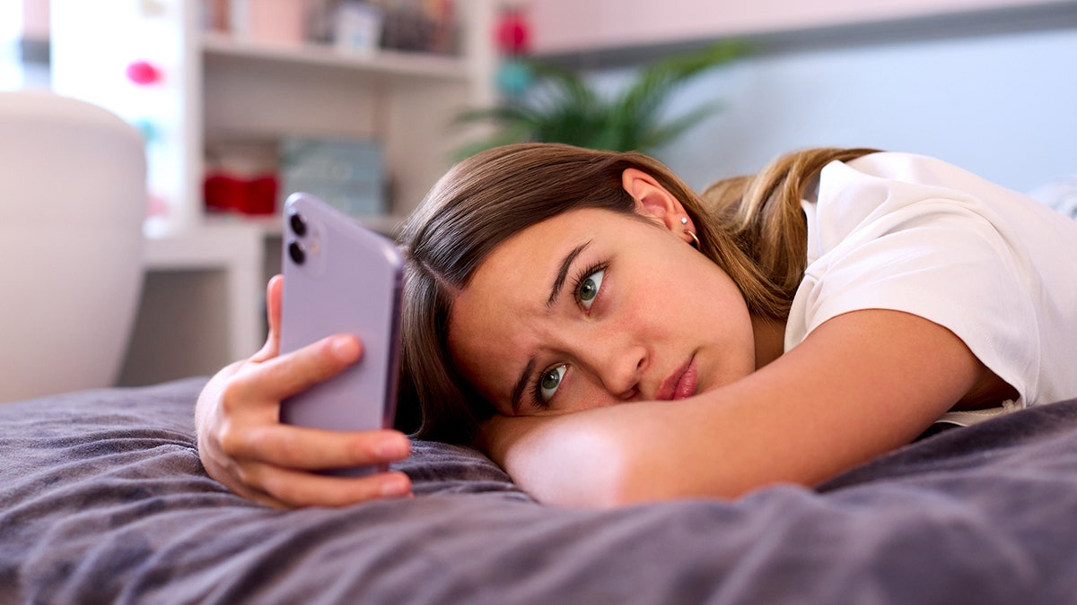 Teenage girl lying on bed at home looking at her cell phone