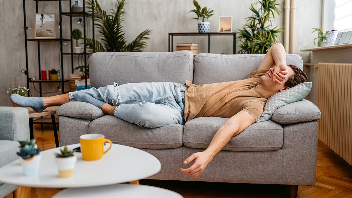 A man is lying on a grey couch. He is wearing blue jeans and a tan T-shirt.