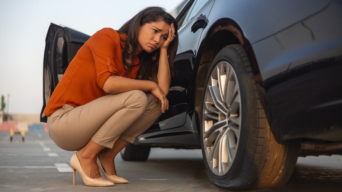 Sad woman with brown hair in an orange blouse and tan pants crouching next to a car with a flat tire.