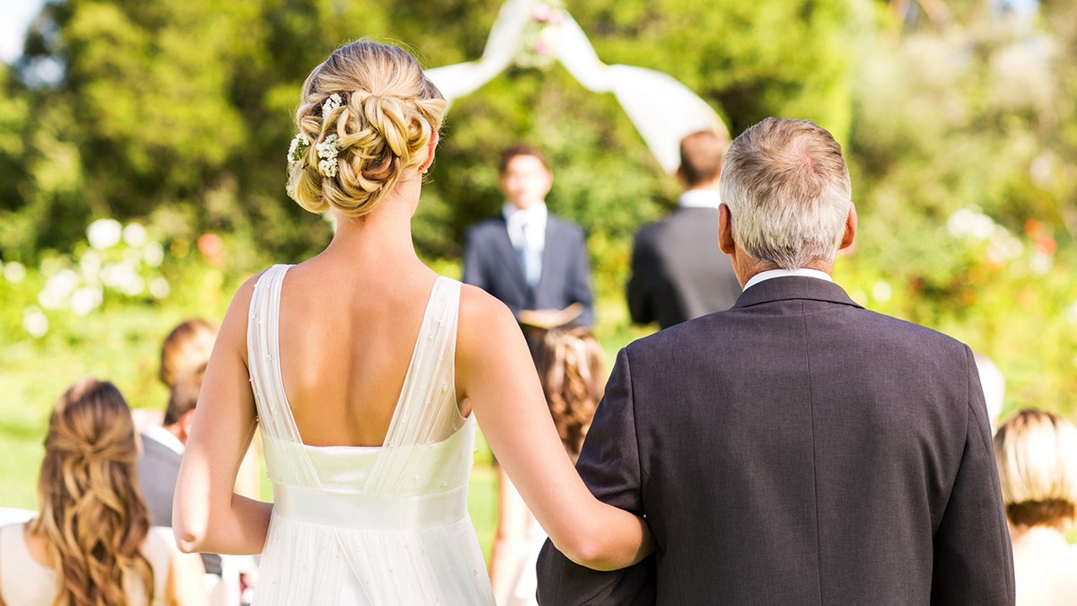 Vista trasera de la novia y el padre caminando hacia el altar con gente al fondo durante una boda al aire libre.