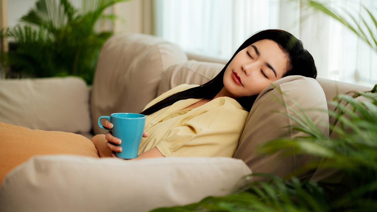 Una mujer relajada se echa una siesta en un sofá, con una taza de café en la mano