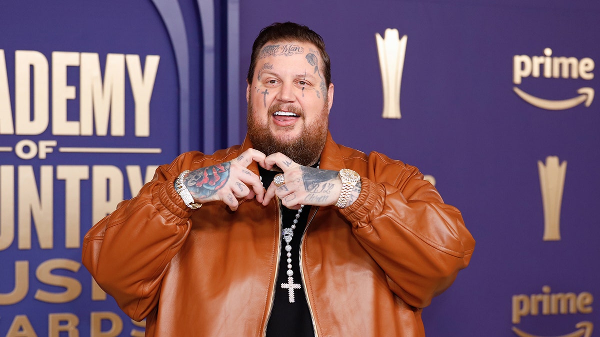 Jelly Roll in a burnt orange leather jacket makes a heart with hands at the ACMS