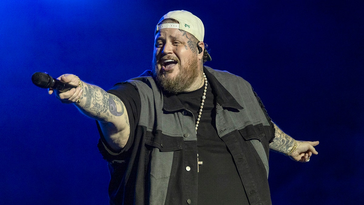 Jelly Roll in a grey and black shirt and backwards hat points microphone outward toward the crowd
