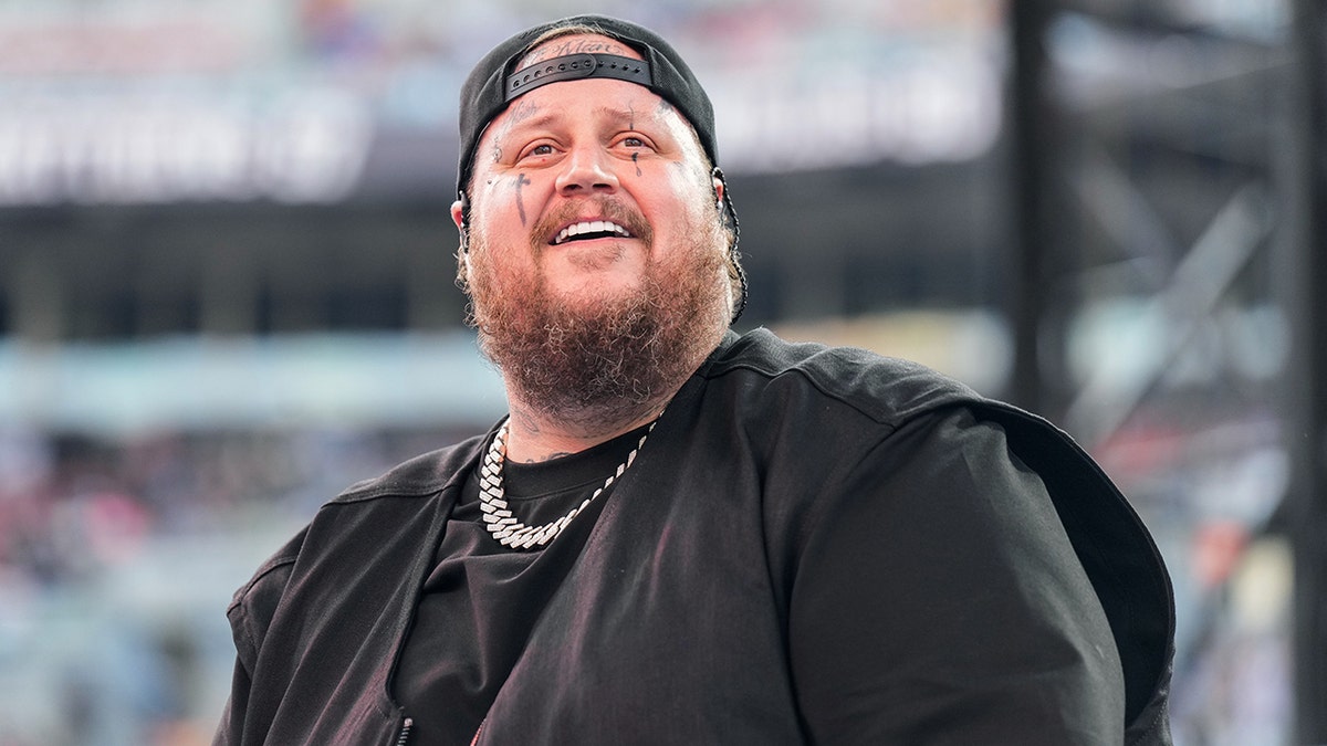 Jelly Roll in a black shirt and black upside-down hat looks up and smiles at the crowd