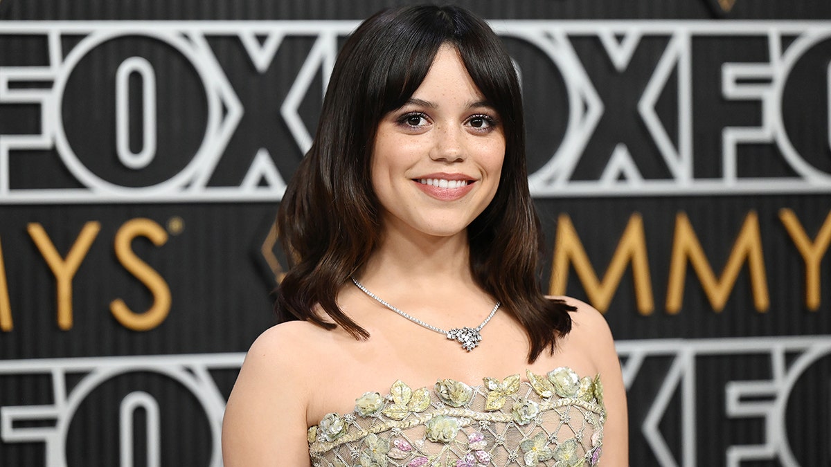 Jenna Ortega em um vestido sem alças sorri no tapete