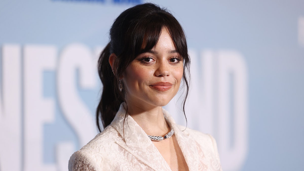 Jenna Ortega sorri suavemente em um blazer branco no tapete