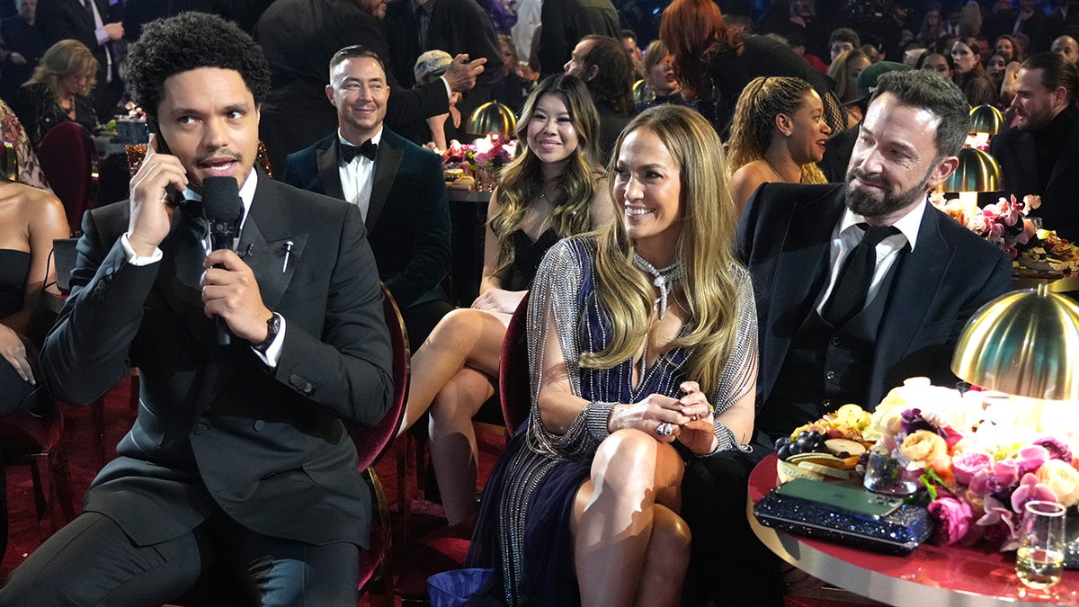 Trevor Noah habla por el micrófono mientras presenta los Grammy mientras Jennifer Lopez, sonriente, y Ben Affleck le miran desde la mesa de al lado