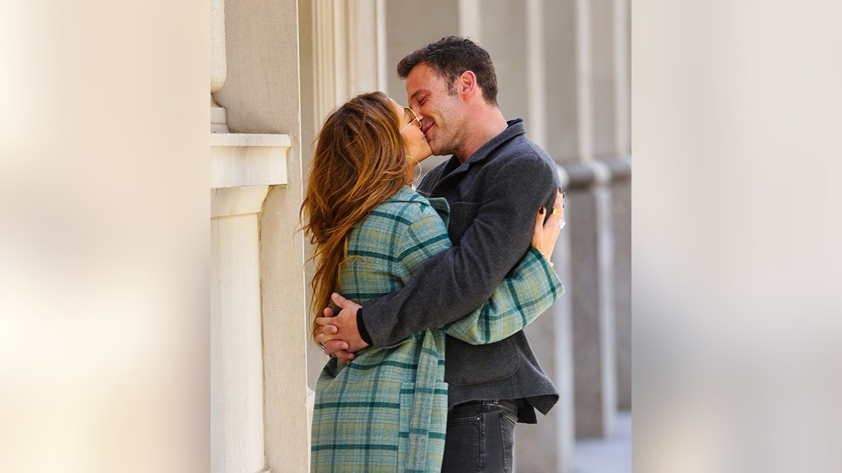 Jennifer Lopez con una chaqueta verde a cuadros besa a un sonriente Ben Affleck que la rodea con sus brazos