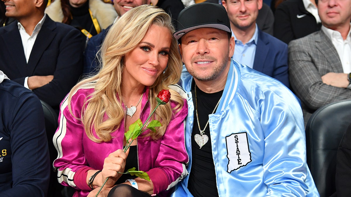 Jenny McCarthy in a pink jacket smiles softly with her husband Donnie Wahlberg in a bright blue jacket