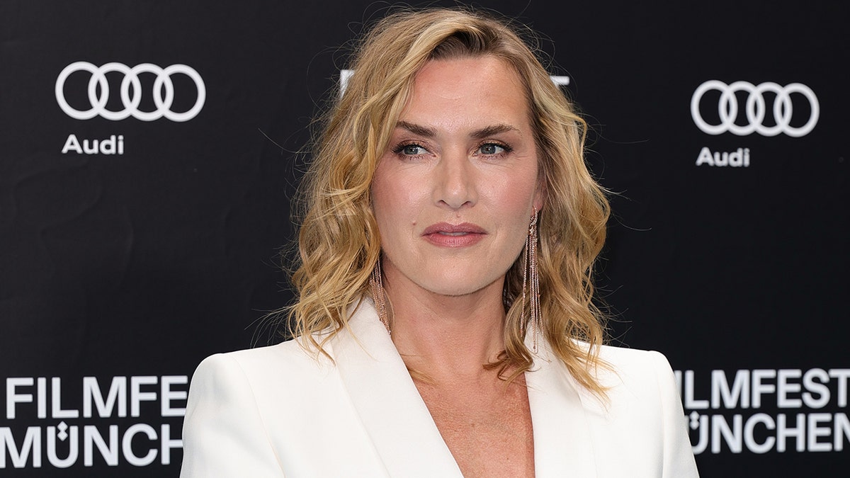 Kate Winslet melihat ke kanannya di atas karpet mengenakan setelan jas putih