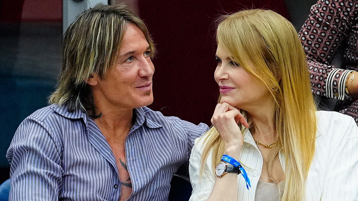 Keith Urban con una camisa de rayas azules y blancas mira hacia arriba mientras Nicole Kidman con la mano bajo la barbilla mira con adoración a su marido en el US Open