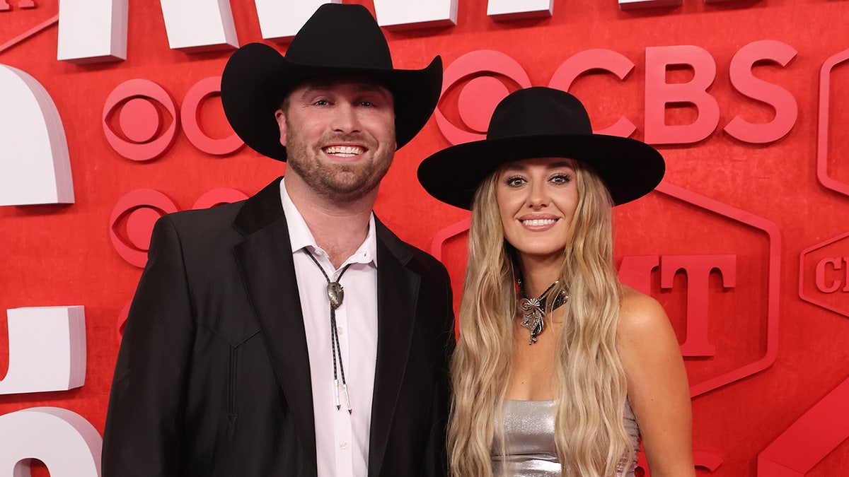 Devlin Hodges in a black suit and cowboy hat smiles with Lainey Wilson in silver top and black hat