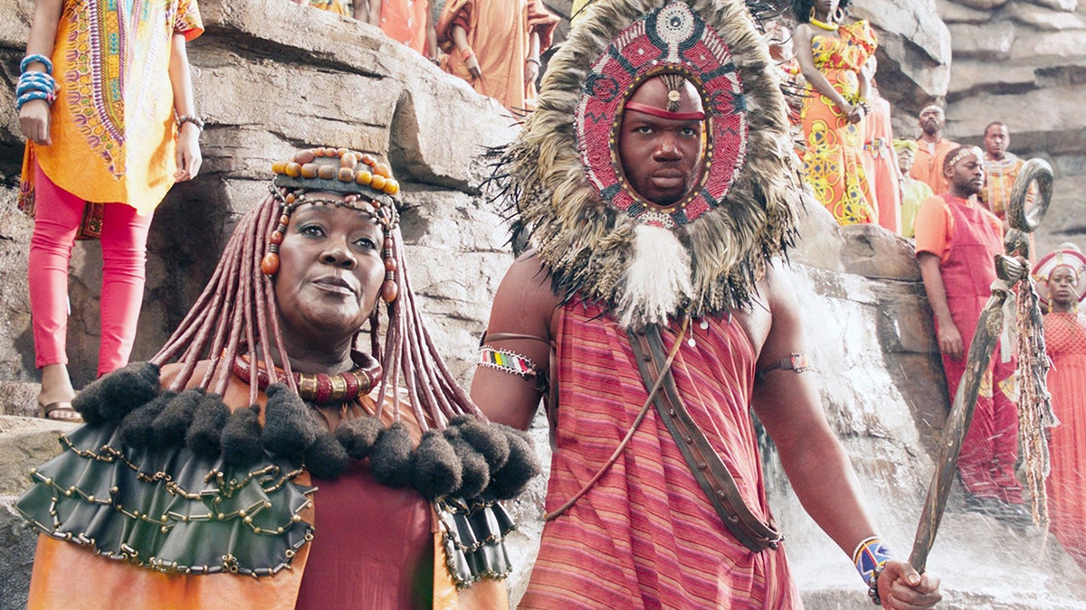 Black Panther scene with Connie Chiume as Zawavari wearing a headpiece and orange cape next to a man in a headdress and red shirt