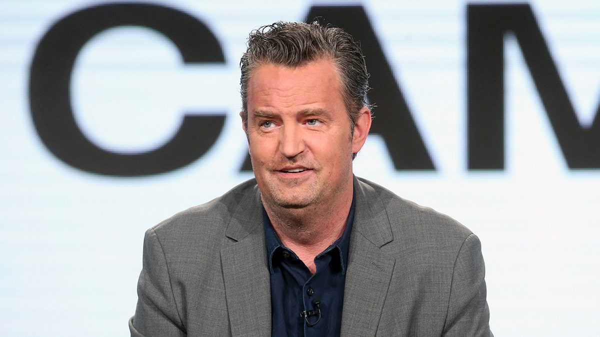 Matthew Perry con traje gris y camisa oscura mira a lo lejos en el escenario