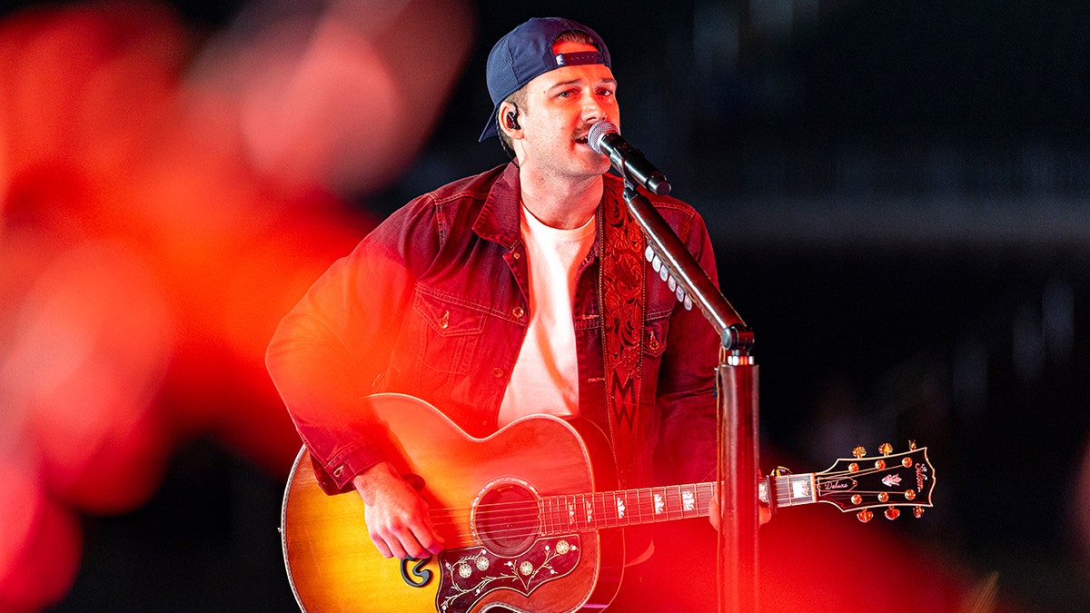 Morgan Wallen in a red shirt and backwards hat performs on stage with a guitar