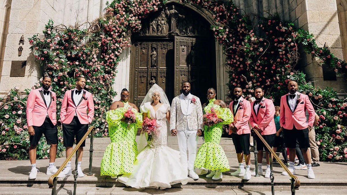 Bride and groom in white, two bridesmaids in lime green polka dot dresses, and groomsmen in pink jackets with black shorts. Everyone in sneakers.