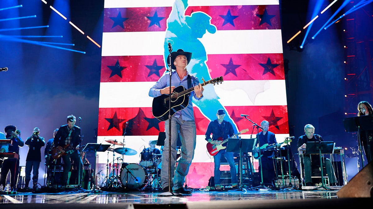 Parker McCollum in a blue shirt and jeans plays guitar on stage for a tribute to Toby Keith