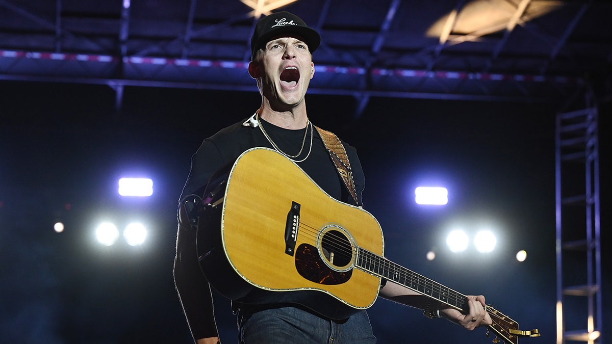 Parker McCollum looks like he's yelling on stage in a black long sleeve shirt with his guitar across his chest