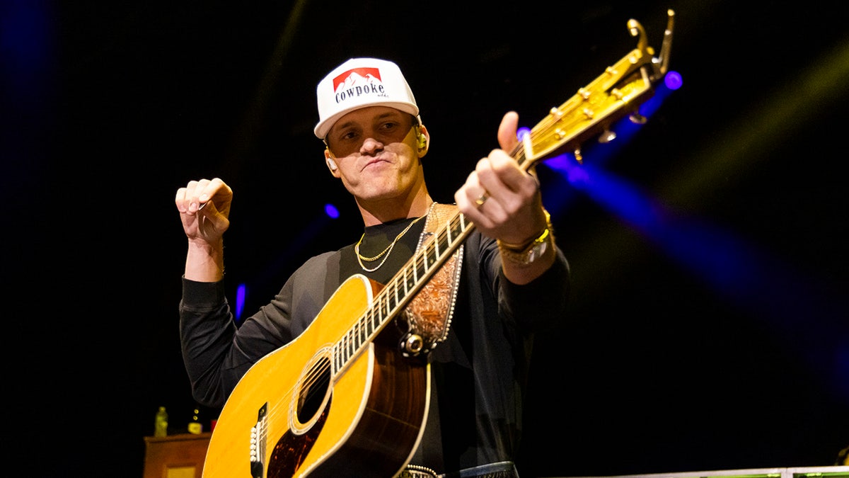 Parker McCollum in a black long sleeve shirt and white hat looks down at his guitar on stage