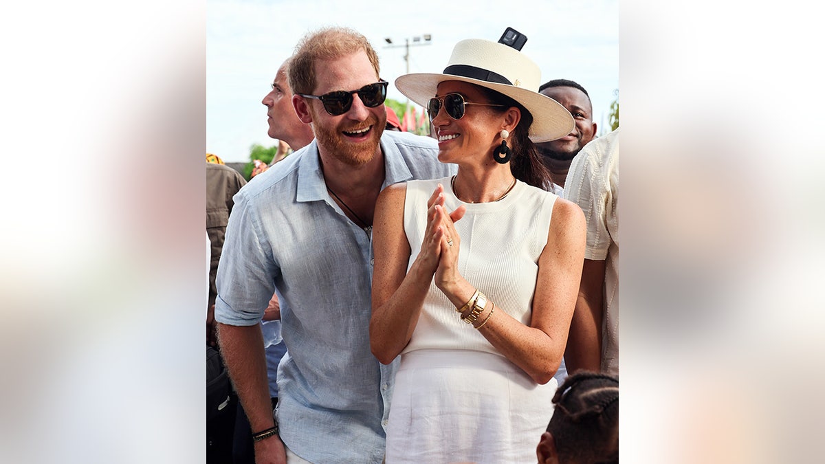 El príncipe Harry se ríe con una camiseta azul y gafas de sol puestas junto a Meghan Markle con un top y un sombrero color crema y una falda blanca