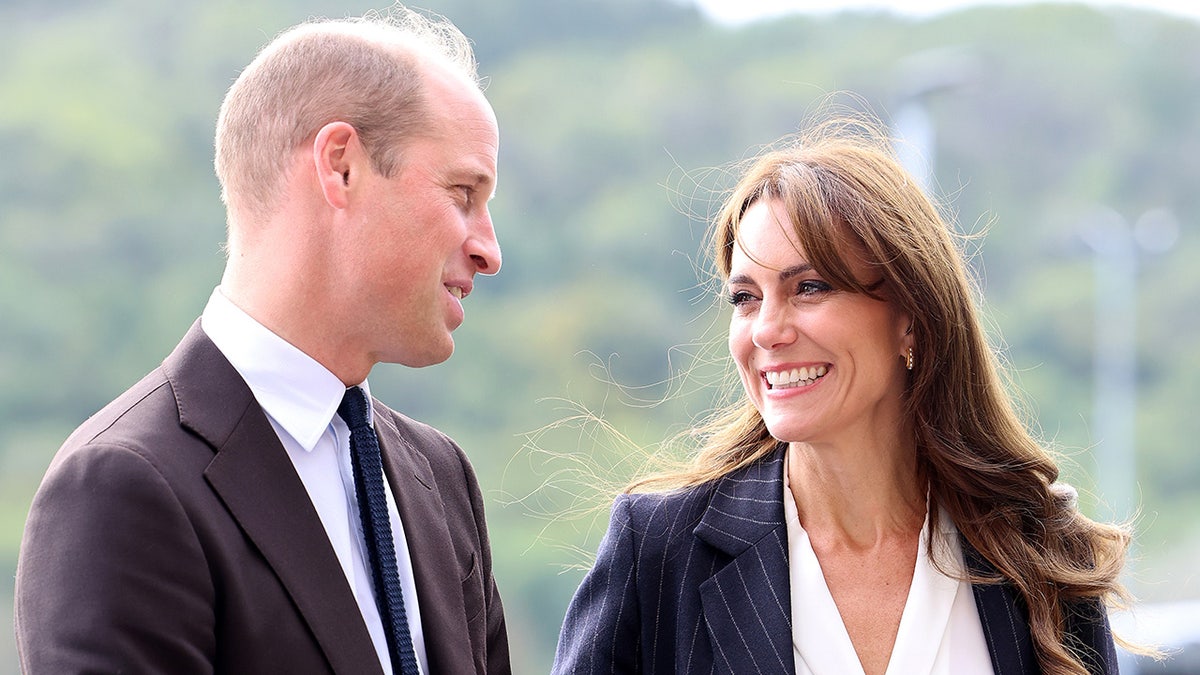 Prince William in a brown suit smiles and looks down at wife Kate Middleton in a blue blazer and white shirt