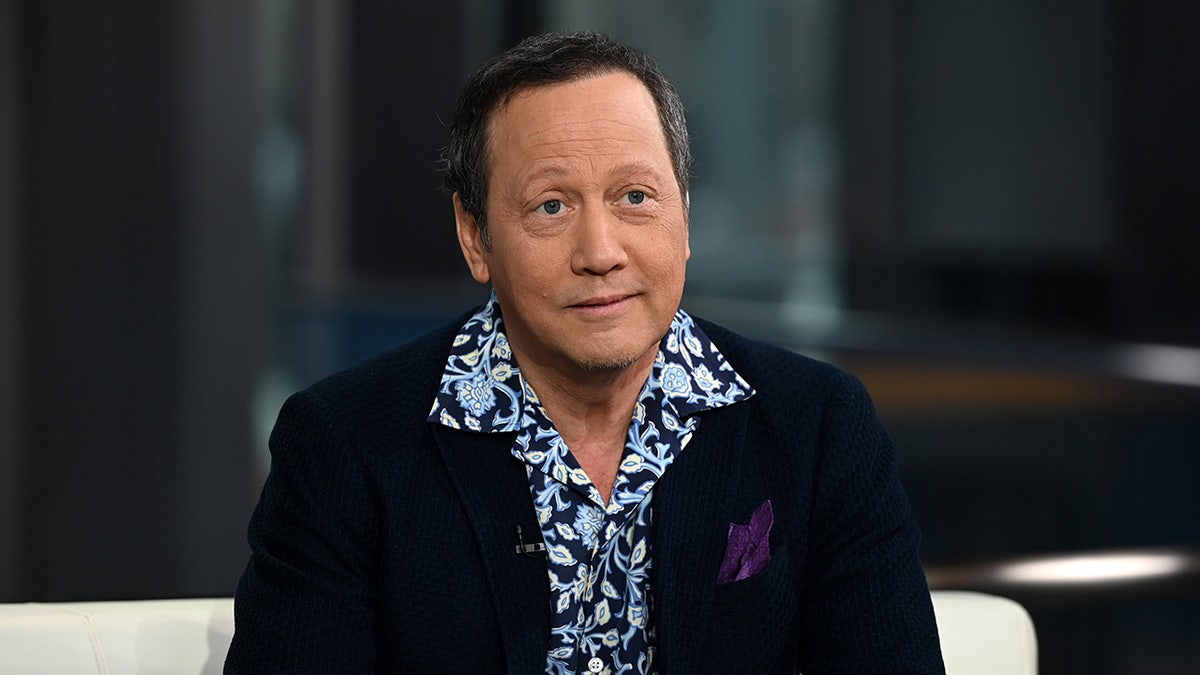 Rob Schneider con una chaqueta azul marino oscura y una camisa azul estampada sentado en una silla