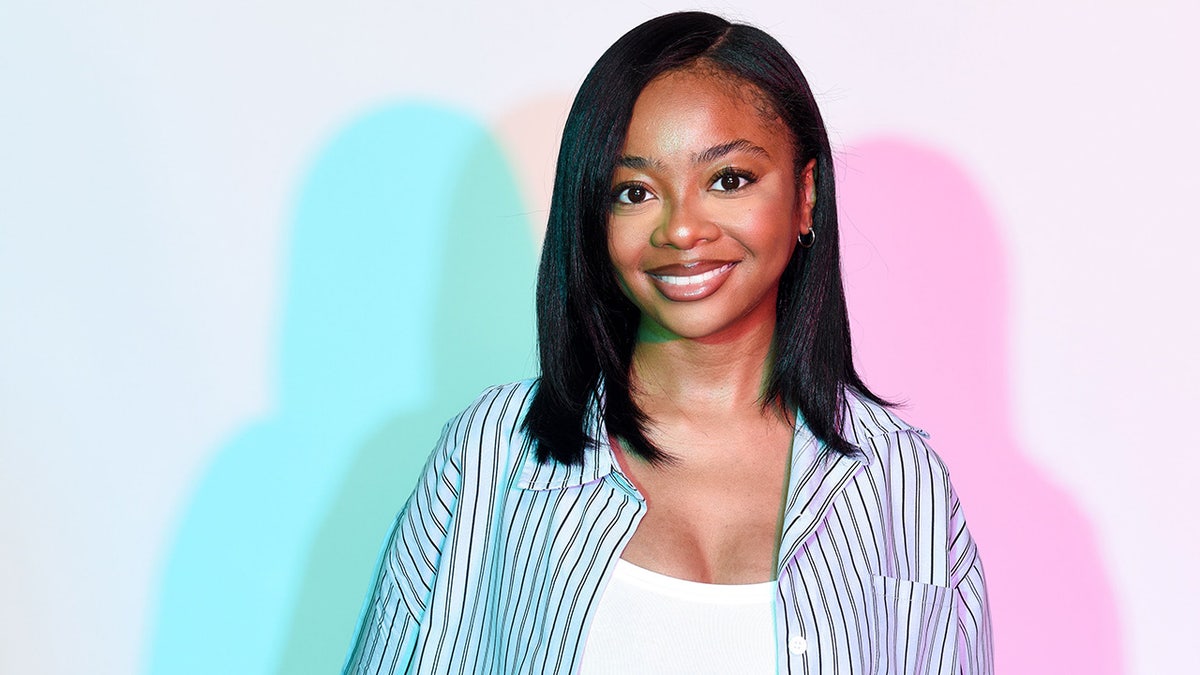 Skai Jackson con chaqueta vaquera y camisa blanca sonríe a la cámara con una sombra rosa y azul de su trasero