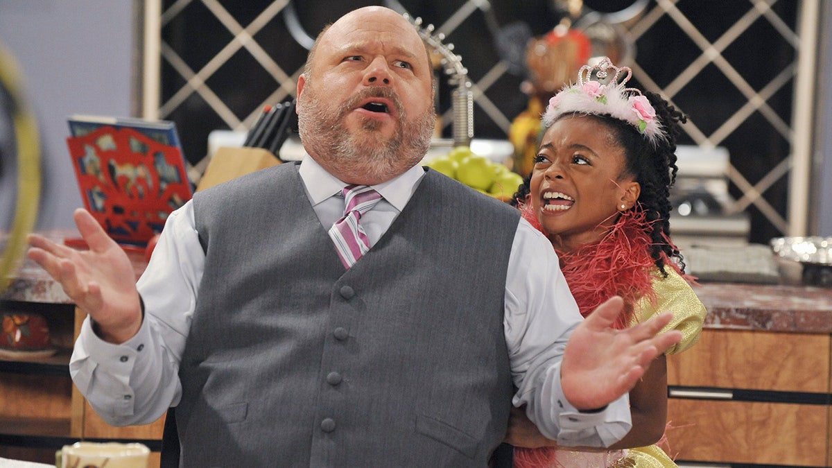 A young Skai Jackson on "Jessie" in a crown looks up at Kevin Chamberlain as Bertram Winkle in a grey sweater vest