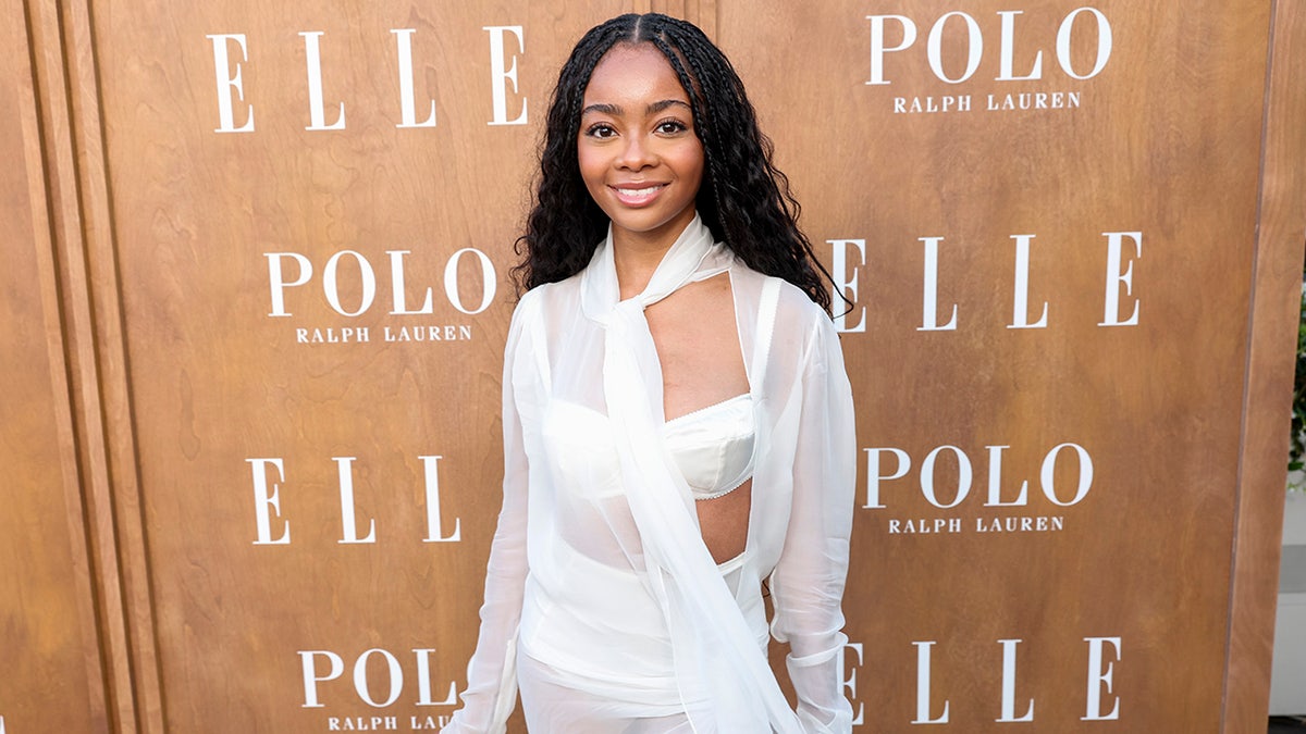 Skai Jackson en sujetador blanco y top blanco transparente sonríe en la alfombra