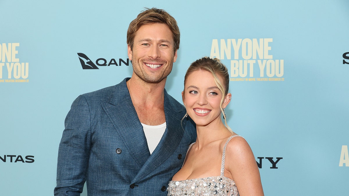 Glen Powell in a blue blazer smiles broadly on the carpet with Sydney6 Sweeney in a light blue/silver beaded dress