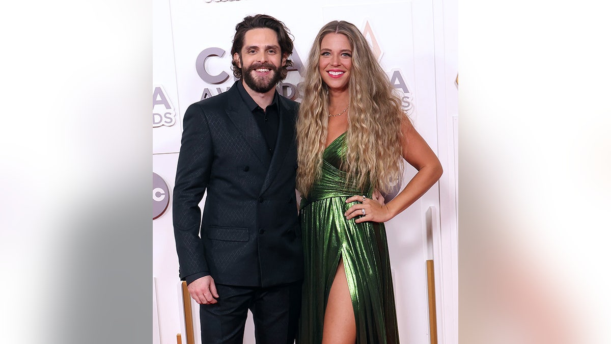 Thomas Rhett em um terno preto sorri com a esposa Lauren com cabelos super longos e cacheados e um vestido verde metálico