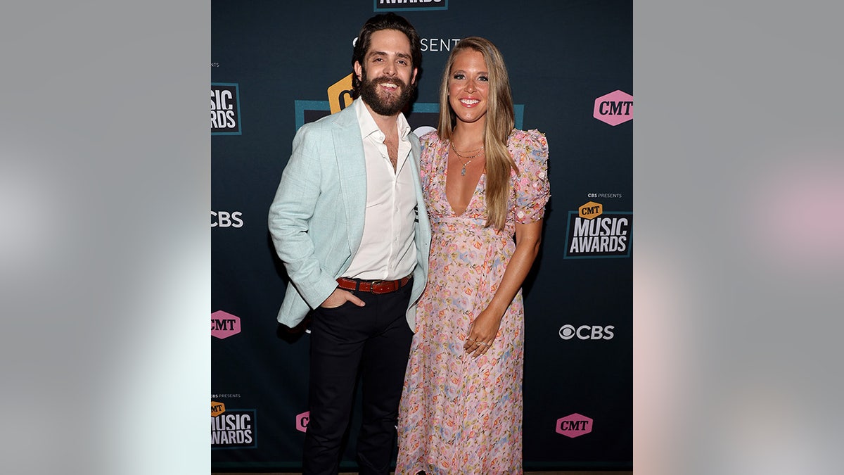 Thomas Rhett sorri em um terno azul gelo ao lado de sua esposa Lauren em um vestido rosa estampado no tapete