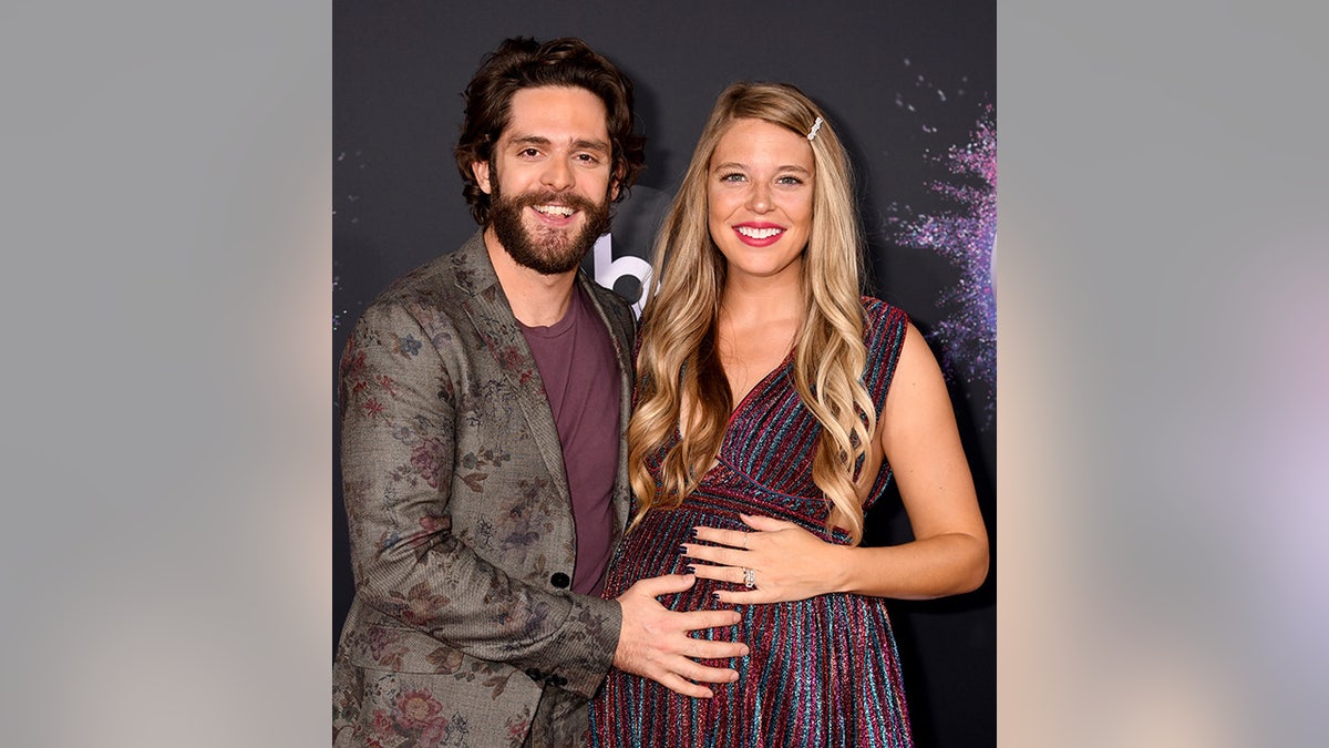 Thomas Rhett in a patterned grey suit and purple shirt puts his hand on wife Lauren's baby bump at the American Music Awards