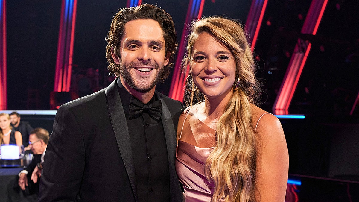 Thomas Rhett in a black suit and shirt smiles with wife Lauren in a satin pink dress at the CMA Awards