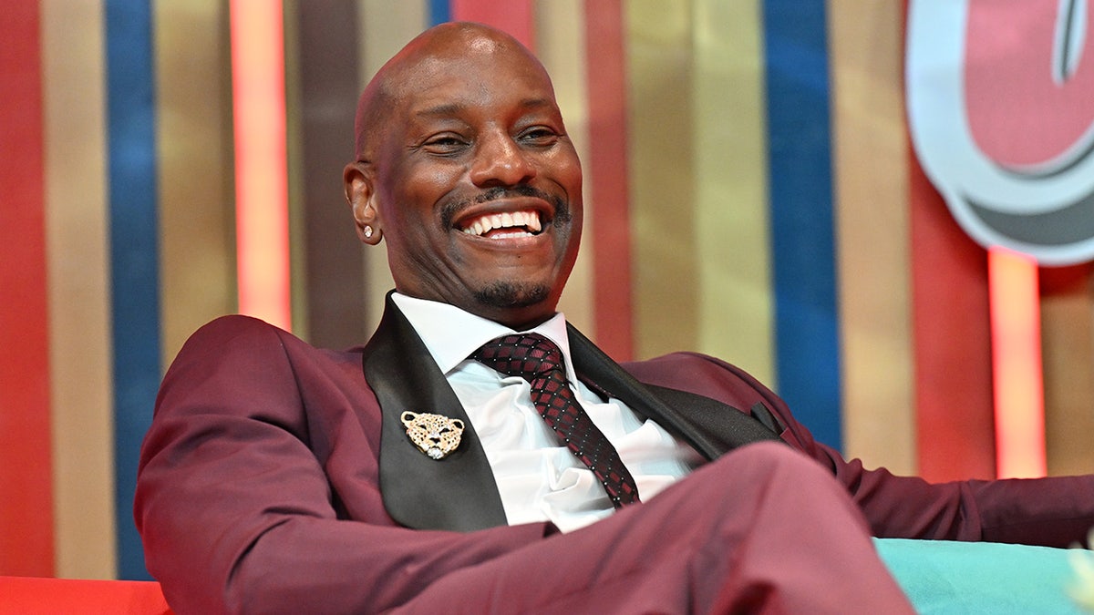 Tyrese Gibson smiles on stage in a burgundy suit