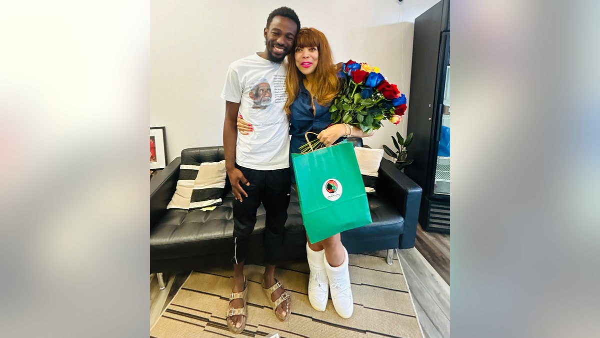 Wendy Williams smiles in a blue outfit, poses with the shop owner, holds a teal bag and wears white moon boots