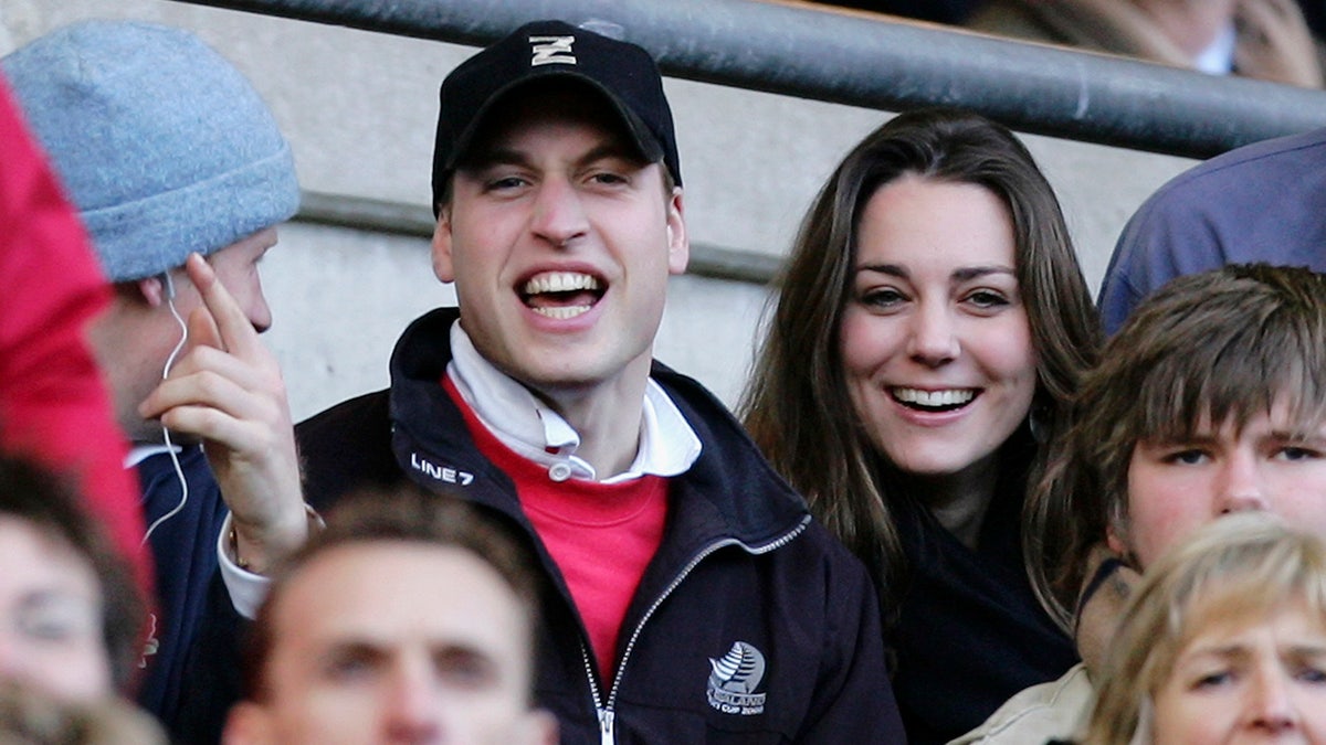 El príncipe Guillermo con sombrero y chaqueta negros y camisa roja se ríe en un partido con Kate Middleton de negro