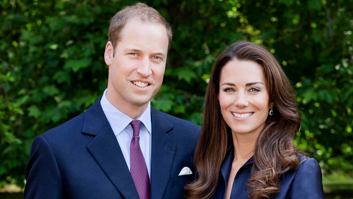 Prince William in a blue blazer and purple tie smiles next to wife Kate Middleton, also in blue