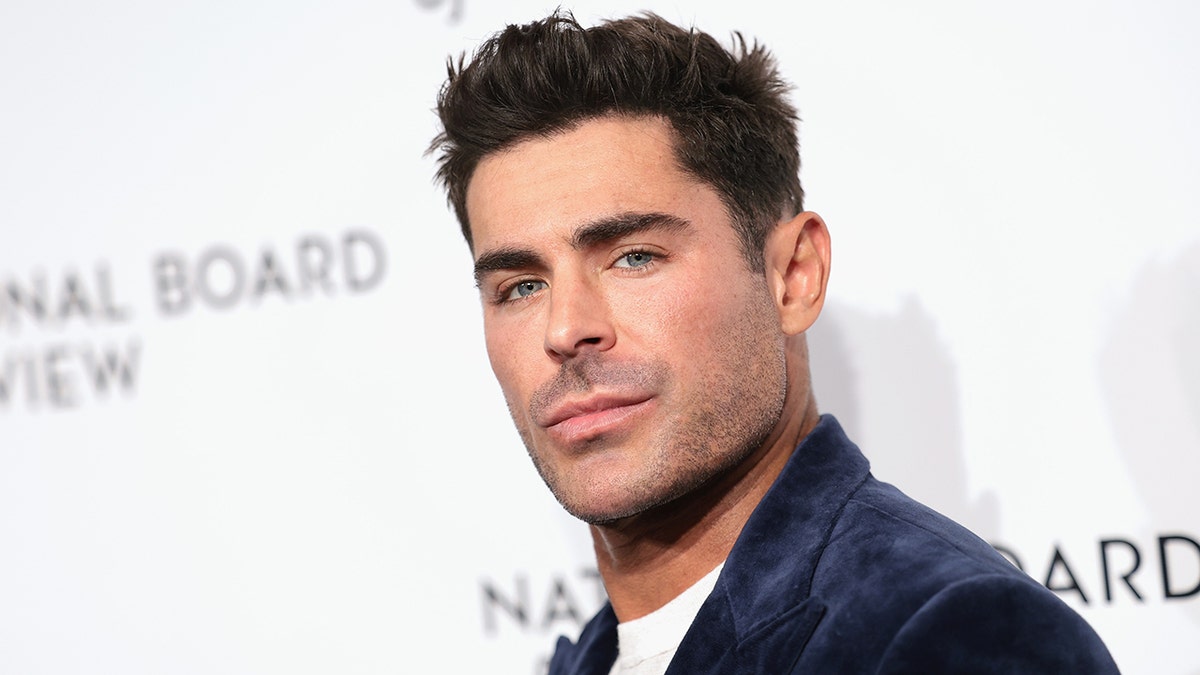 Zac Efron terlihat sedikit menoleh ke belakang ke kamera dalam balutan setelan beludru biru tua di atas karpet