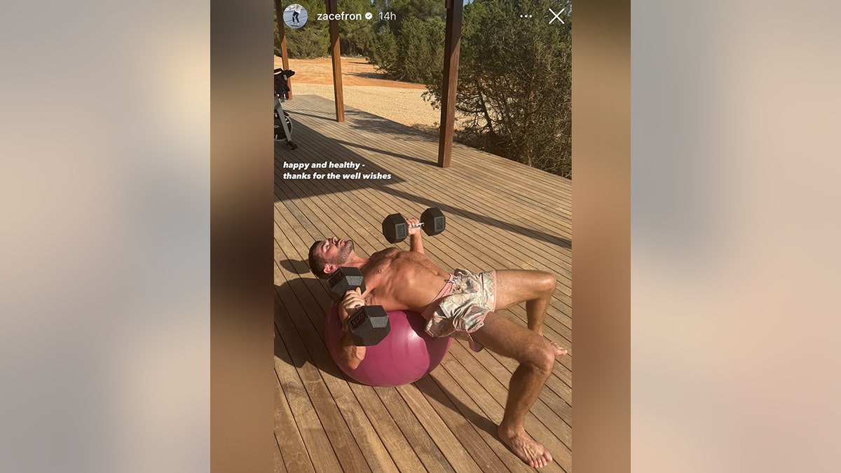 Zac Efron lying back on a pink ball lifting weights shirtless