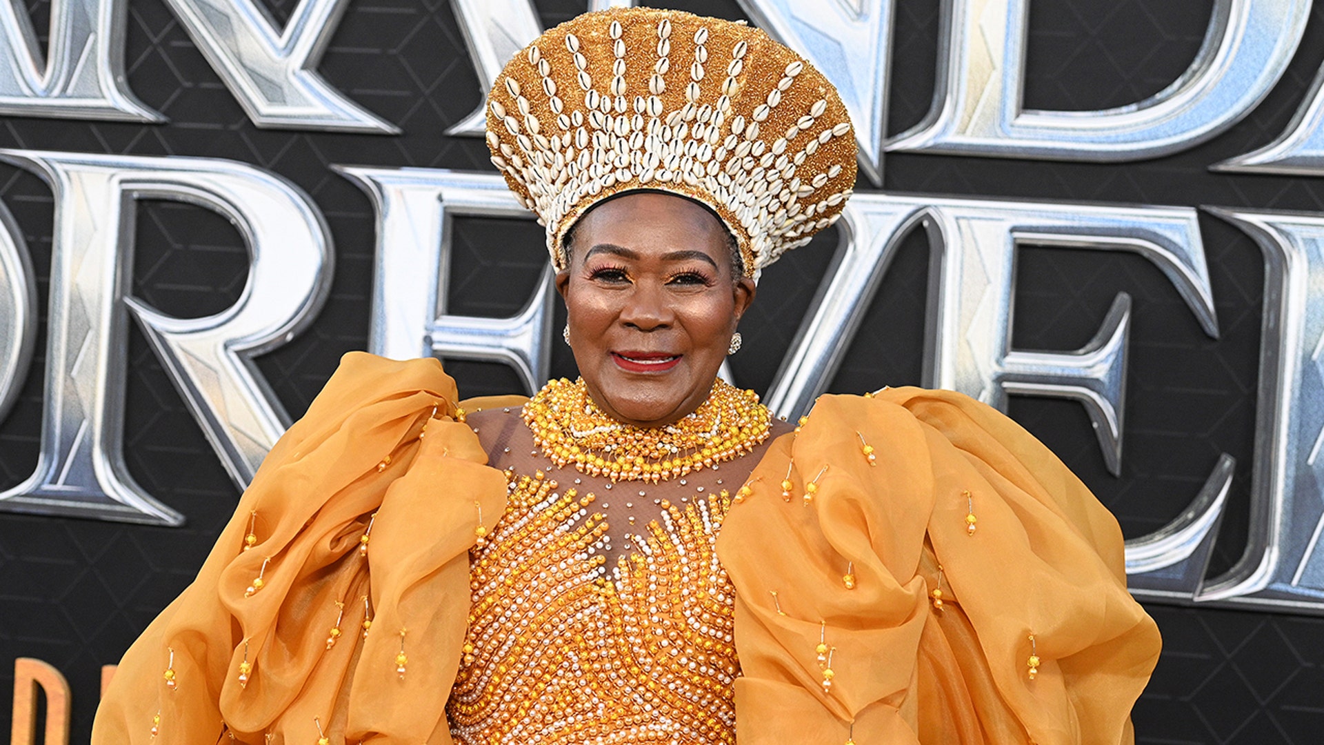 Connie Chiume in a golden yellow dress with puffy sleeves and a matching head piece at the "Wakanda Forever" premiere