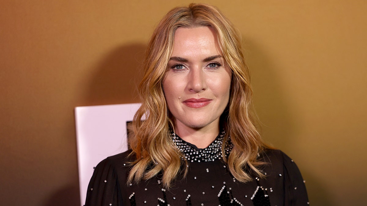 Kate Winslet dalam balutan gaun hitam dengan sedikit hiasan menatap kamera di atas karpet