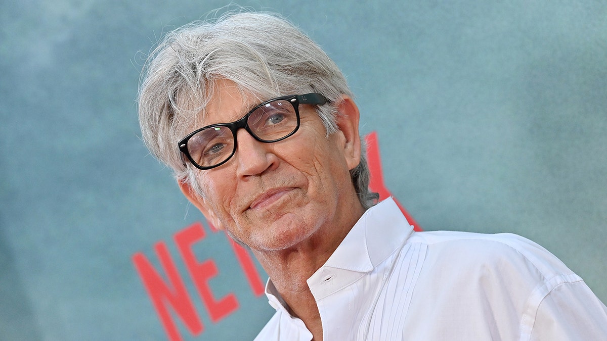 Eric Roberts wears black glasses and a white shirt on the carpet