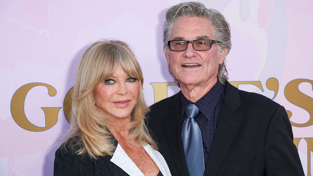 Goldie Hawn con un blazer negro brillante con solapas blancas mira a su derecha en la alfombra junto a Kurt Russell con traje negro y corbata azul